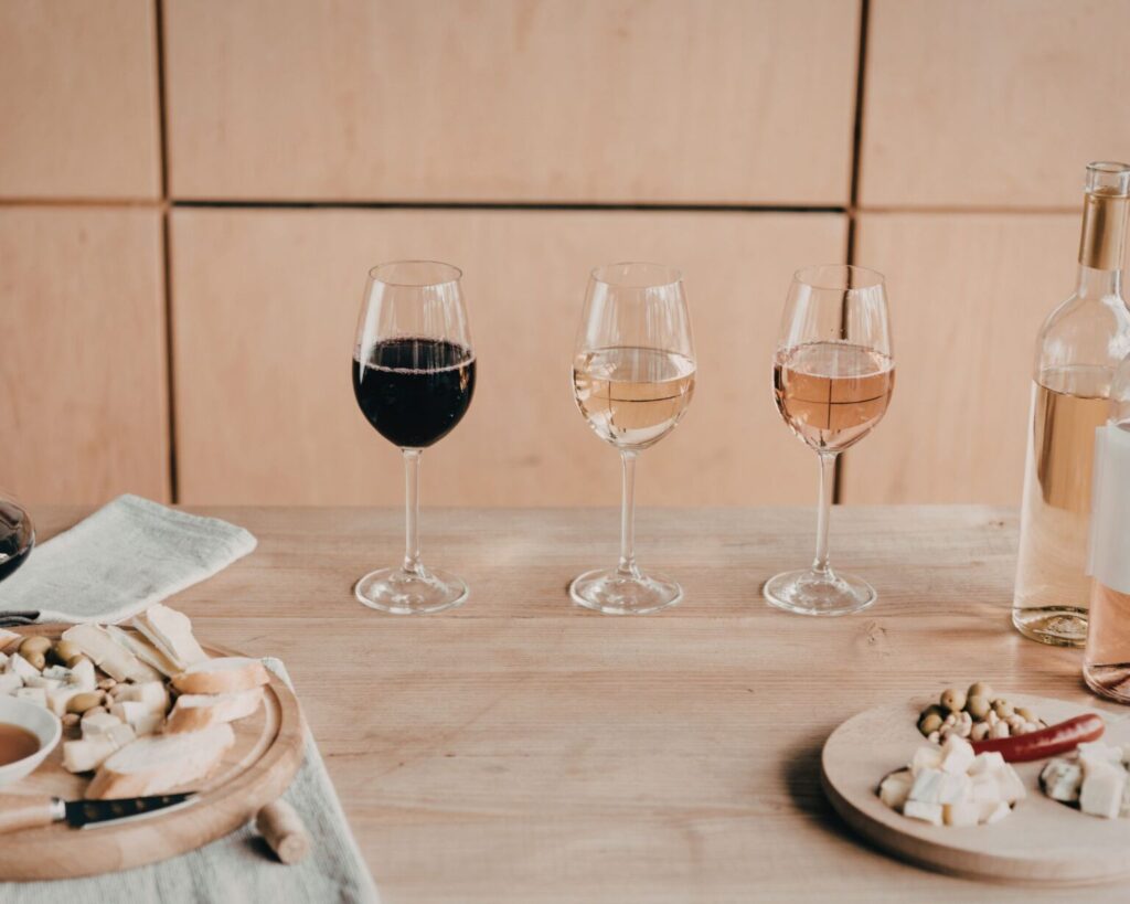 Three wine glasses are lined up on a light wooden table, containing red wine, white wine, and rosé wine, respectively. A partially visible bottle of wine and two small wooden boards with cheese, olives, and sliced baguette are also on the table, set against a light wood paneled wall.