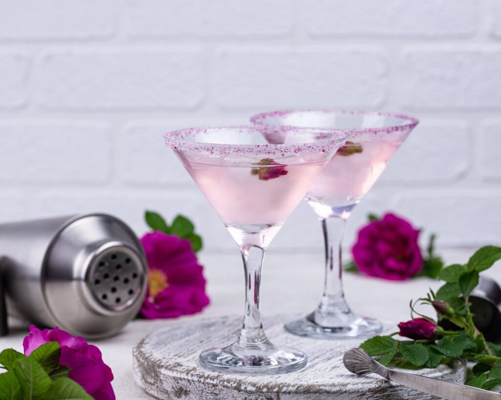 Two elegant martini glasses filled with a light pink cocktail, such as a Rose Cosmo, with sugar-rimmed edges and garnished with small dried rosebuds. The glasses are set on a white wooden serving board next to a cocktail shaker and fresh magenta wild roses, against a white brick wall.
