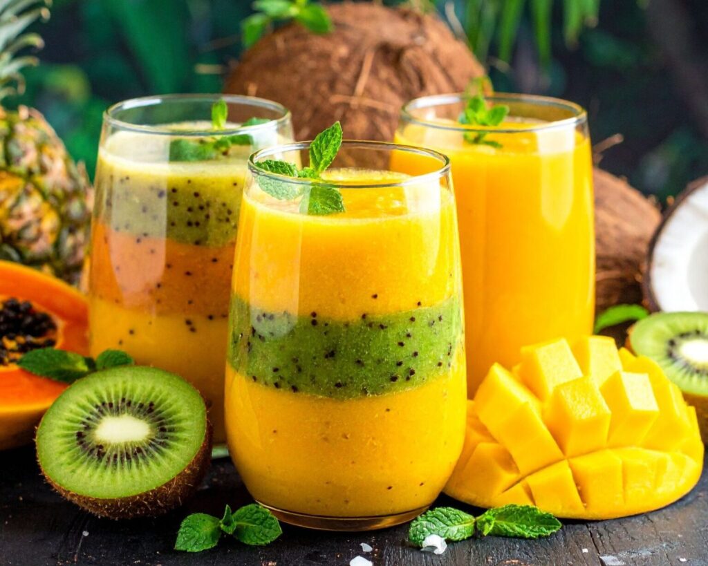 Three clear glasses filled with layered tropical fruit smoothies or juices, featuring alternating layers of bright yellow (mango or pineapple) and vibrant green (kiwi), garnished with mint leaves. The drinks are surrounded by fresh fruit, including halved papaya, sliced kiwi, coconut, and sliced mango, with lush green tropical foliage in the background.