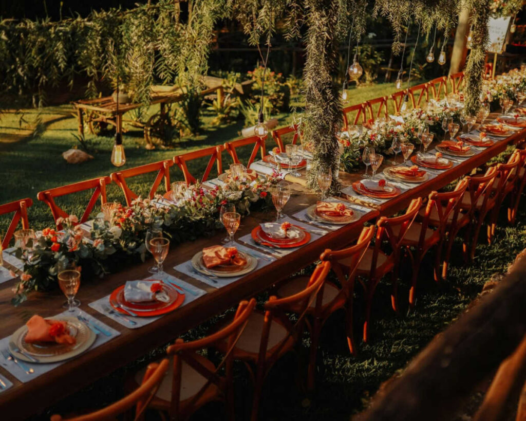 A long wooden table set for an outdoor event, lined with cross-back wooden chairs. The table is elegantly set with white and red plates, orange linen napkins, and a floral runner featuring white and orange flowers and greenery. Edison-bulb string lights hang over the table, with the setting sun casting a warm glow on the scene.