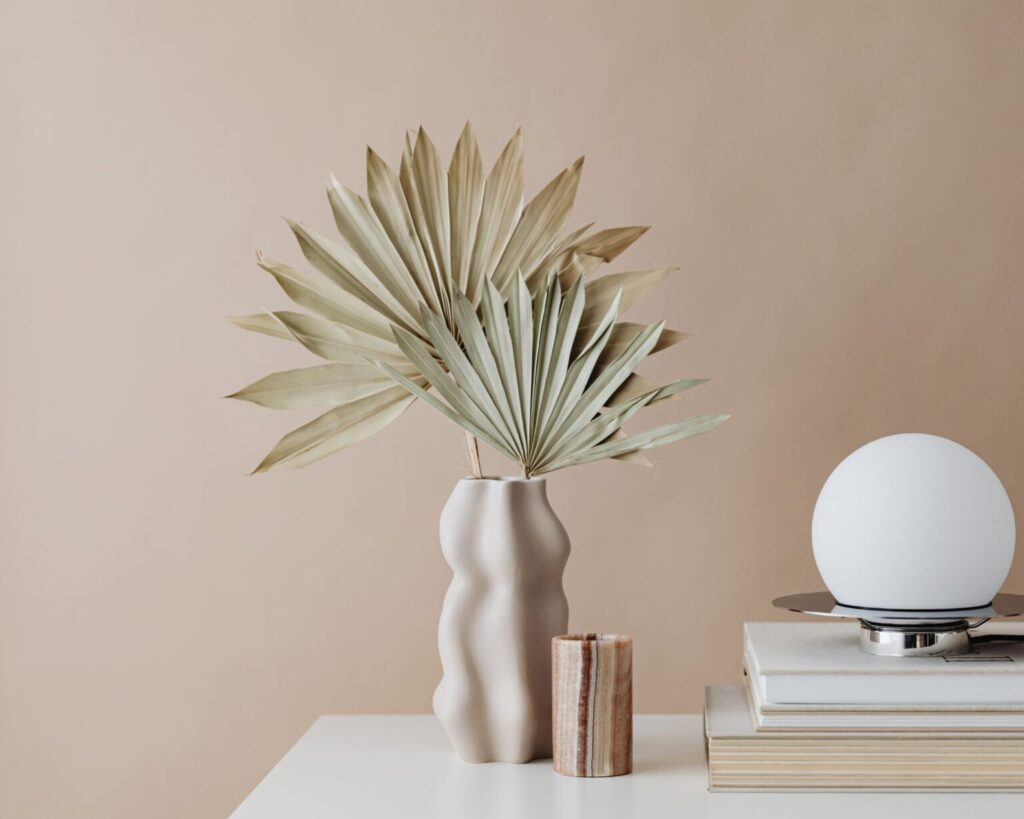 A minimalist centerpiece arrangement featuring a large, dried palm frond in a textured, wavy beige ceramic vase, set next to a brown striped onyx candle holder. The arrangement is placed on a white surface next to a stack of books topped with a modern, spherical white lamp, all against a pale tan background.