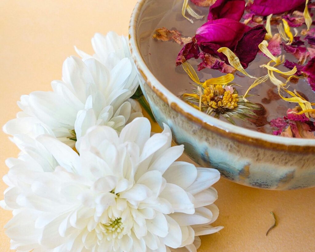 A close-up of a DIY centerpiece featuring two large, white chrysanthemum blossoms next to a rustic ceramic bowl filled with water and floating dried petals in shades of pink, magenta, and yellow. The setup is on a pale peach or yellow background.