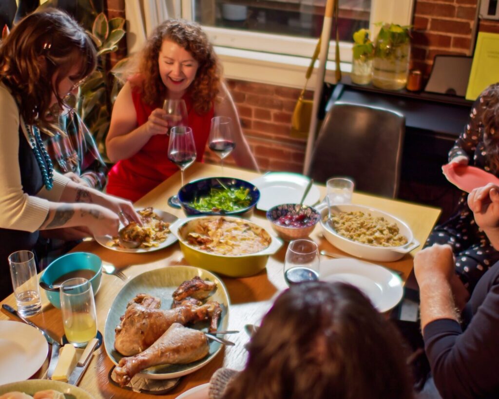 A group of friends gathered around a wooden table indoors for a casual dinner, which includes roast turkey legs or chicken, a casserole, a green salad, and other side dishes. The woman in the center in a red dress is laughing while holding a wine glass, and another person is serving food onto a plate.
