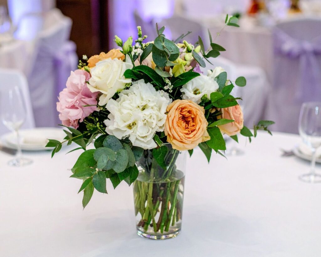 A centerpiece bouquet in a clear glass vase, featuring a mix of white, pale pink, and peach roses and white carnations or hydrangeas, with eucalyptus and other greenery. The arrangement is placed on a white-linen covered table in a banquet hall with draped chairs in the background.