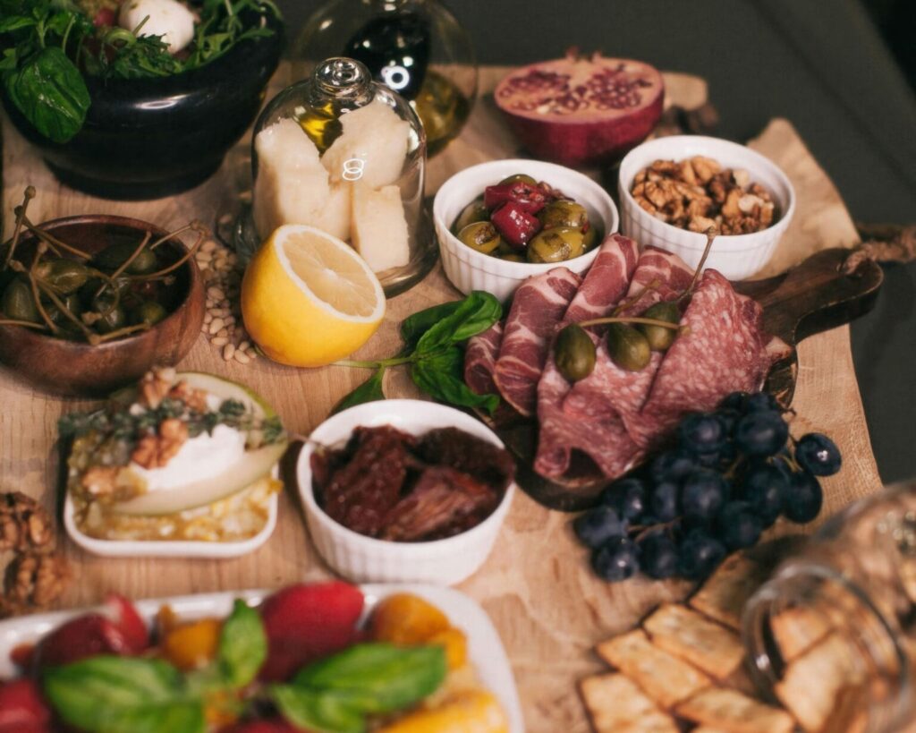 A rustic charcuterie and antipasto board featuring folded slices of cured meats (salami and ham), mixed olives, walnuts, sun-dried tomatoes, a wedge of lemon, capers, fresh mozzarella under a glass cloche, grapes, crackers, and a caprese salad in a black bowl.