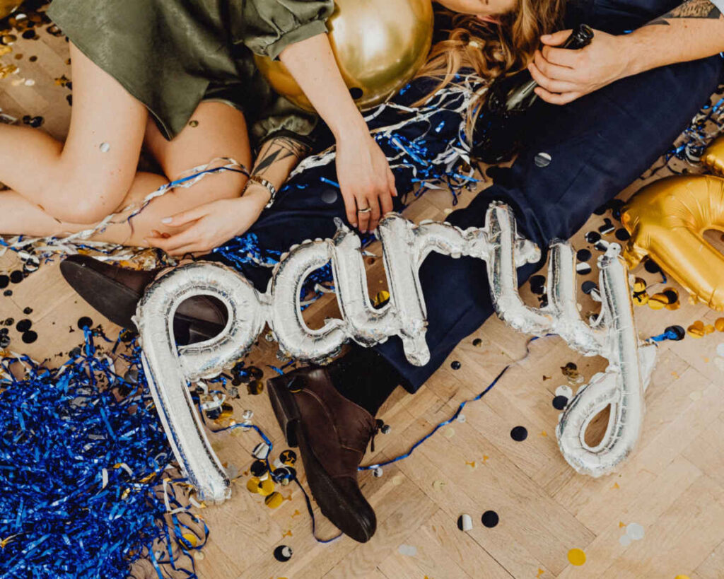 A close-up of a man and woman sitting or lying on a light wooden floor after a party, surrounded by silver, gold, and blue confetti and streamers. A silver foil balloon spelling "PARTY" is draped across their legs and the floor.