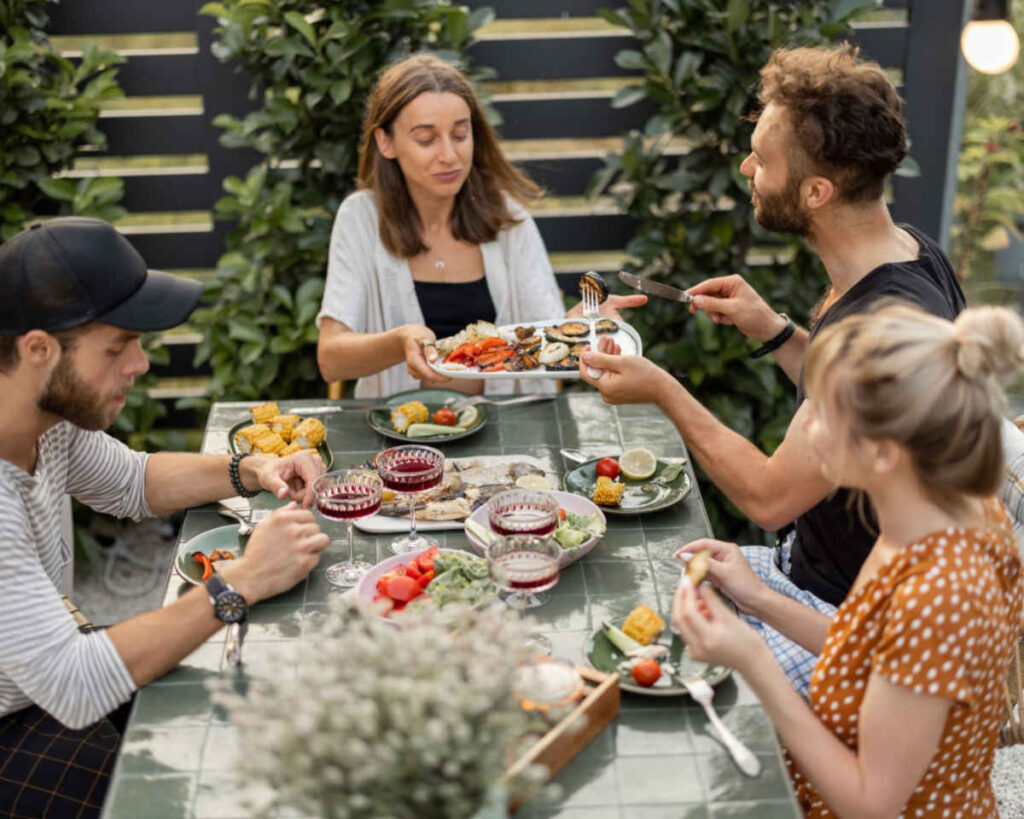 A group of four friends enjoying an outdoor summer dinner party on a patio, sitting around a green tiled table. The table is set with plates of grilled seafood and vegetables, wine glasses, corn on the cob, and cornbread or fritters. One woman is passing a platter of grilled food across the table.