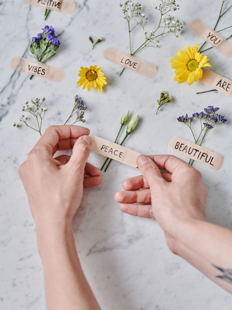 Hands arranging flowers with handwritten affirmation words like peace, love, and beautiful on a marble surface.