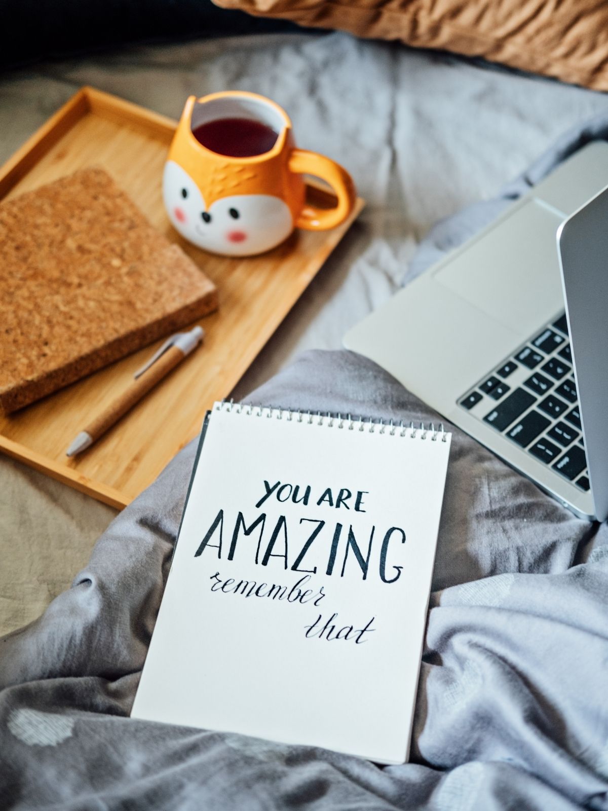Notebook with the handwritten affirmation ‘You are AMAZING, remember that’ on a bed, next to a laptop, a cork notebook, a pen, and a fox-shaped mug with tea.