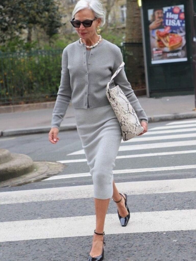 A very stylish older woman with short gray hair is walking across a crosswalk, wearing a chic, matching gray knit set (a fitted cardigan and pencil skirt). She accessorizes with a pearl necklace, bold earrings, cat-eye sunglasses, black patent leather ankle-strap heels, and a large snakeskin print handbag.