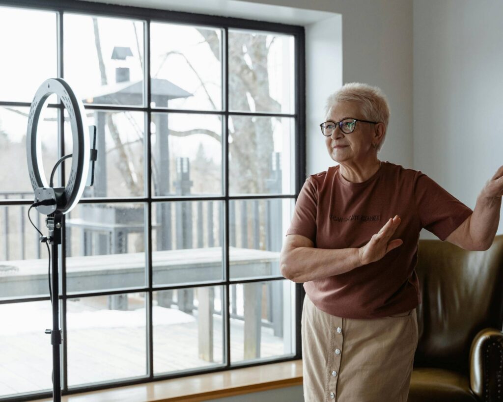 A mature woman with short blonde hair and glasses, wearing a rust-colored t-shirt and a light tan skirt, stands near a large window with a ring light set up on a stand, gesturing with her hands as if recording a video.