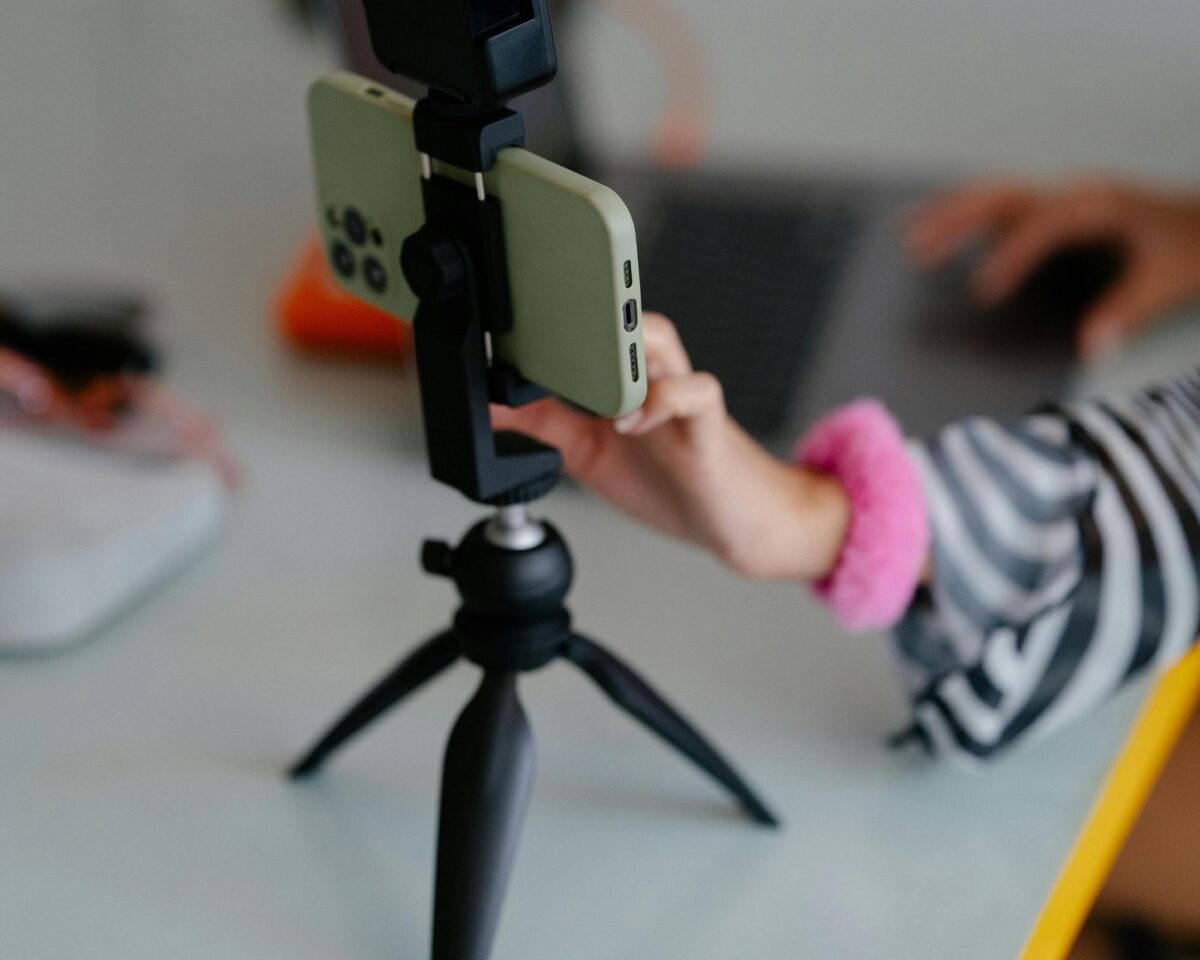 A close-up shot of a smartphone in a pale green case mounted on a small black tripod on a light-colored desk with a yellow edge. In the background, a person wearing a striped shirt and a bright pink wristband uses a laptop, with their hand partially visible near the phone.