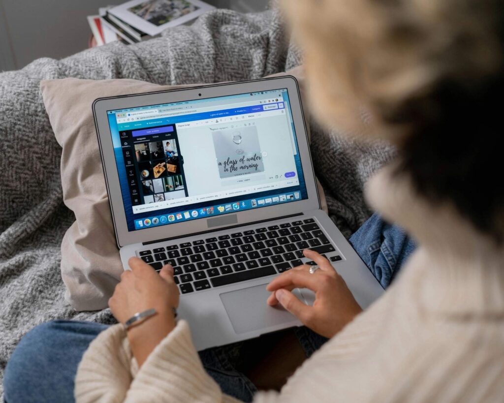 A person, viewed from behind and over the shoulder, sits comfortably on a couch with a gray blanket and cushion, working on a silver laptop. The screen shows the interface of a graphic design program, displaying templates and an image being edited, which includes the text "a glass of water in the morning."