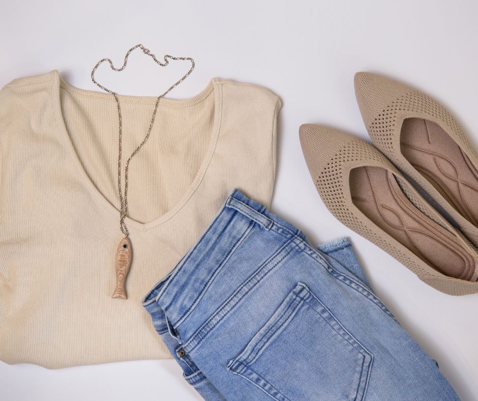 A casual outfit laid flat on a white surface, featuring light wash jeans, a cream V-neck top, tan knit ballet flats, and a silver necklace with a fish pendant.