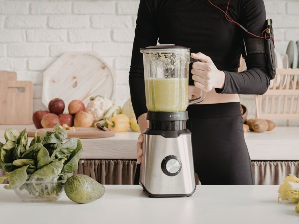 Person in black activewear blending a green smoothie in a blender on a white counter.