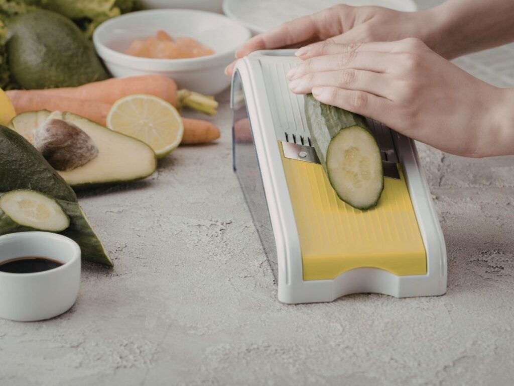 Hands slicing a cucumber with a yellow and white mandoline slicer, surrounded by vegetables.