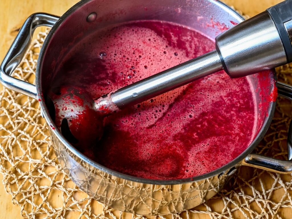 Immersion blender pureeing a bright red liquid or soup in a metal pot.