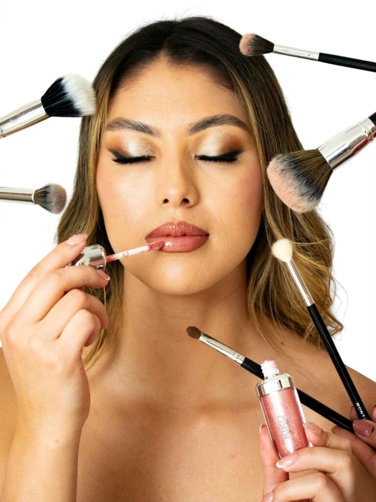 Close-up of woman surrounded by makeup brushes applying lip gloss.
