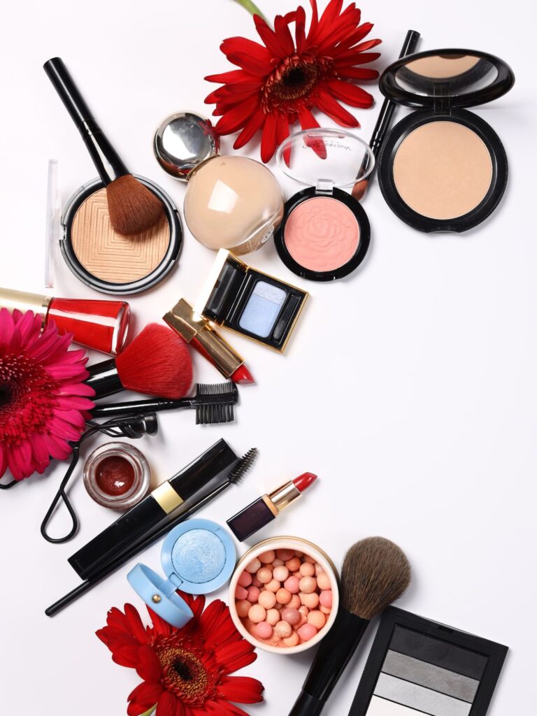 Flatlay of makeup products and brushes with red flowers on a white background.