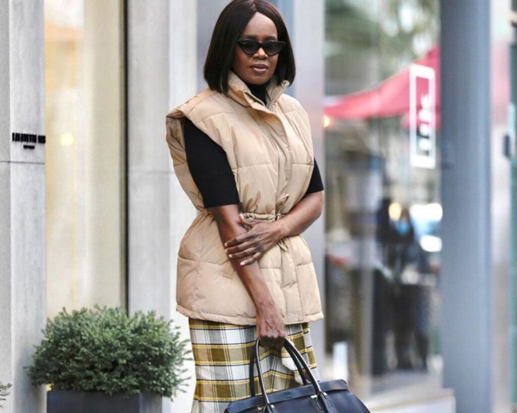 A stylish woman with dark bobbed hair stands outdoors, wearing a beige quilted puffer vest cinched at the waist over a black turtleneck and a long yellow and green plaid skirt. She is wearing sunglasses and carrying a large black handbag.