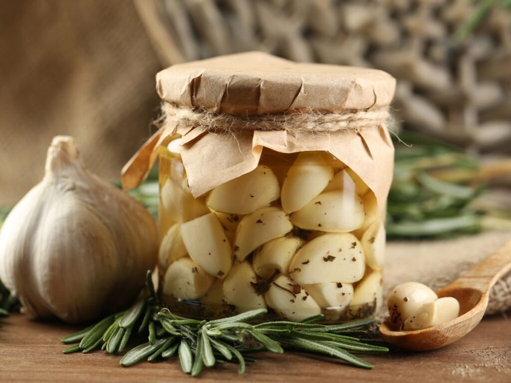 Glass jar of peeled garlic cloves preserved in olive oil, next to a fresh garlic bulb and rosemary.