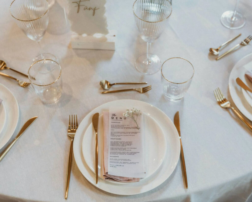 An elegant place setting at a wedding or formal event featuring a white plate layered with a pink napkin and a printed menu card, garnished with a small sprig of baby's breath. The table is set with gold silverware and clear glassware with gold rims, sitting on a white linen tablecloth.