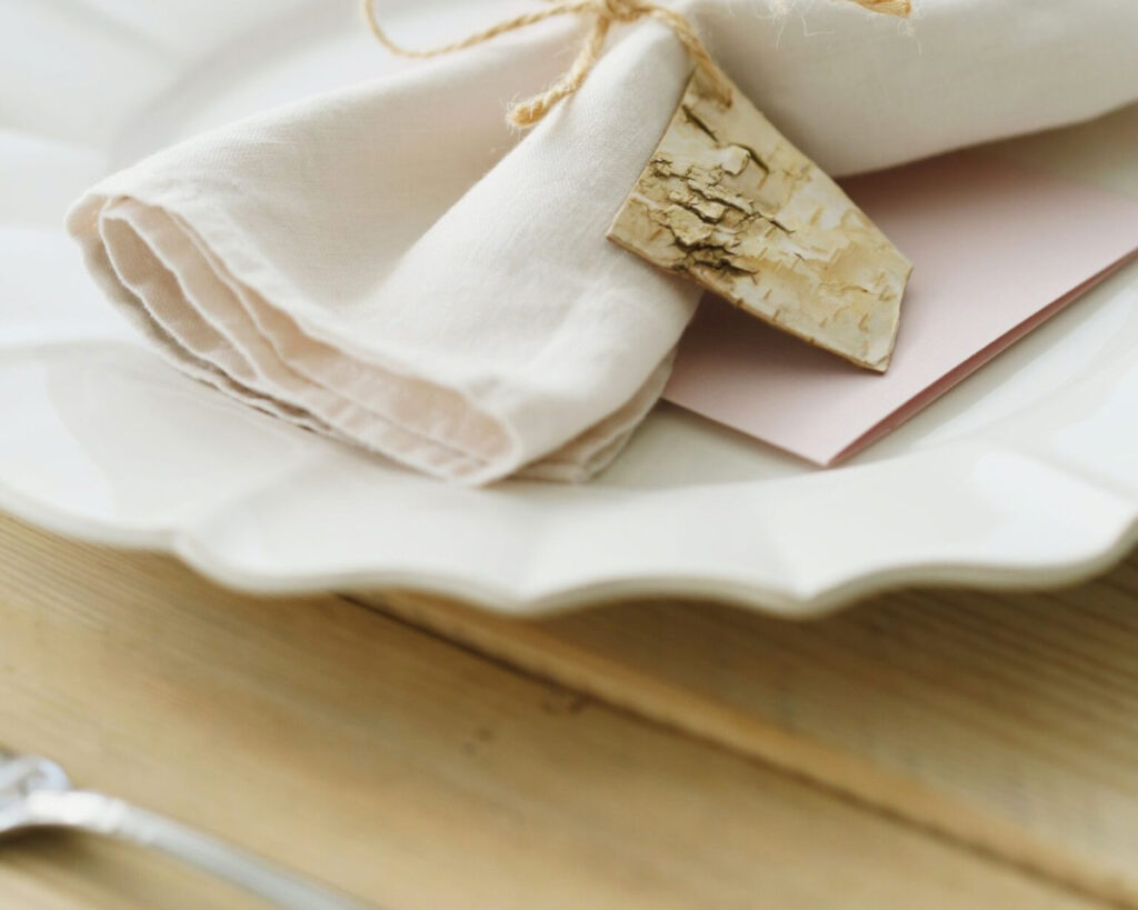 A close-up of a rustic and elegant table setting in soft tones, featuring a folded or rolled light beige linen napkin tied with twine, a natural birch bark tag, and a pale pink envelope or card, all resting on a white ruffled or scalloped dinner plate. The setting is on a light wooden table.