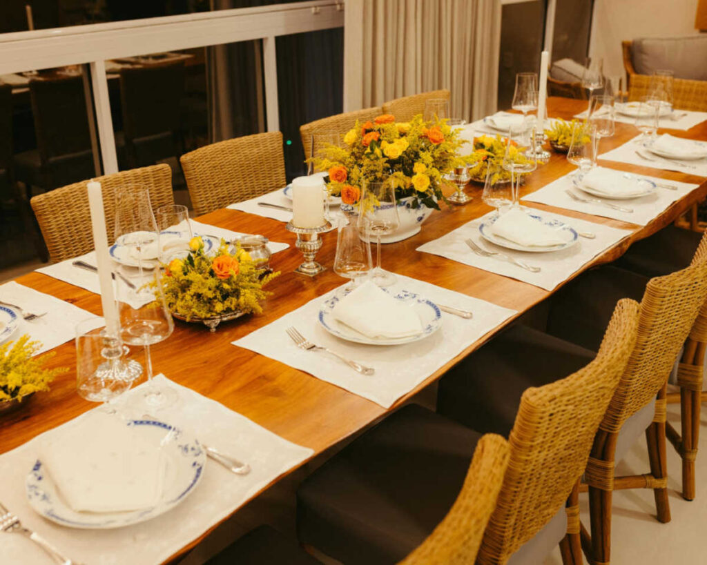 A long, polished wooden dining table set for a dinner party with multiple place settings. The table features woven wicker chairs and white placemats with blue and white patterned plates, decorated with three centerpieces of yellow and orange flowers, white pillar candles, and elegant glassware.