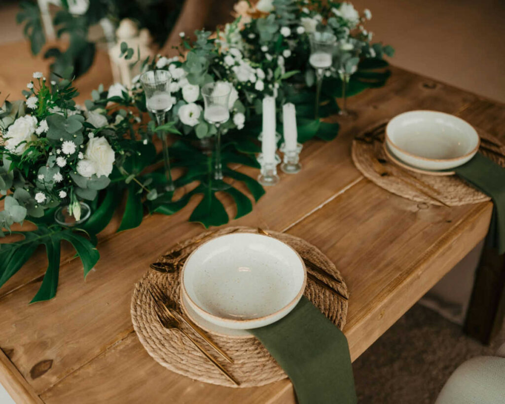 A rustic wooden table set for a tropical or bohemian style event, featuring two place settings with woven round placemats, white speckled bowls, and dark green linen napkins. The table is decorated with a lush floral and greenery runner that includes white roses and large monstera leaves, along with glass candle holders.