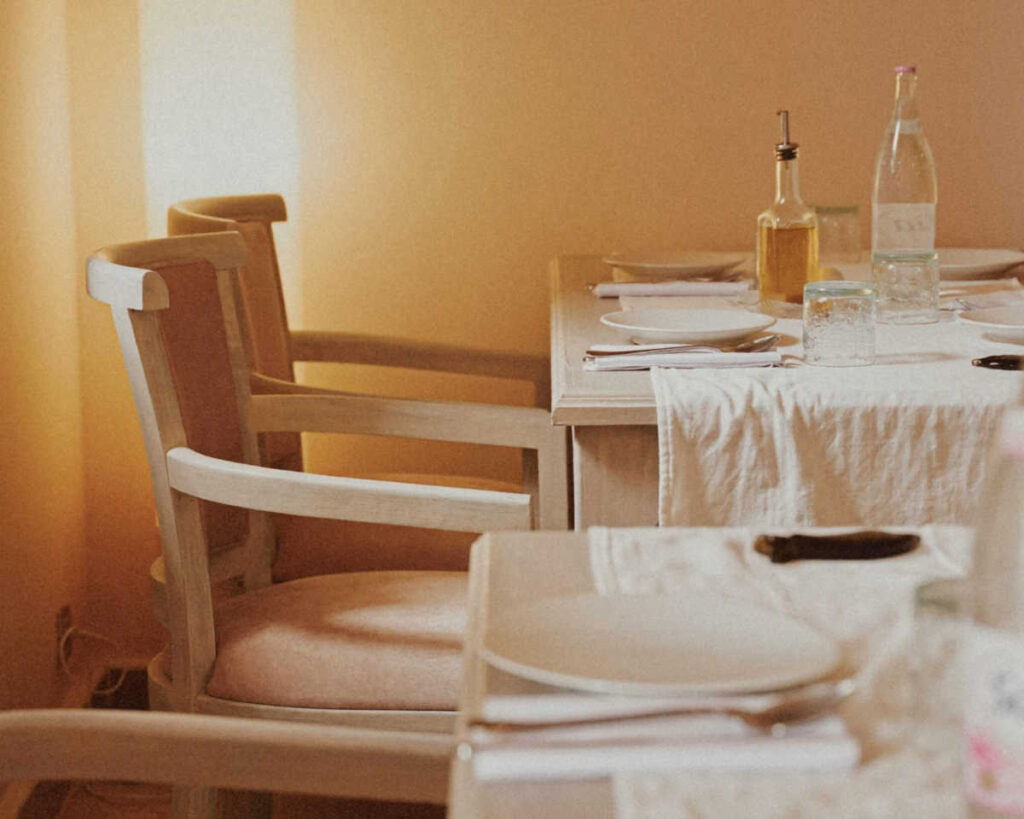 A close-up of a dining table setting with a soft, warm, golden light filtering in. Two light-colored wooden armchairs with pale pink upholstered seats are visible on the left, next to a table set with white plates, a natural linen tablecloth, a bottle of olive oil, and glasses.