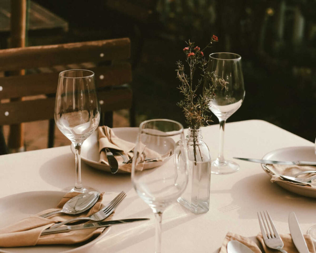 A close-up of a casual outdoor table setting with warm lighting, featuring white plates, silverware wrapped in beige linen napkins, and wine glasses. A small glass bottle serves as a vase for a delicate sprig of red flowers, with a dark wooden chair visible in the background.
