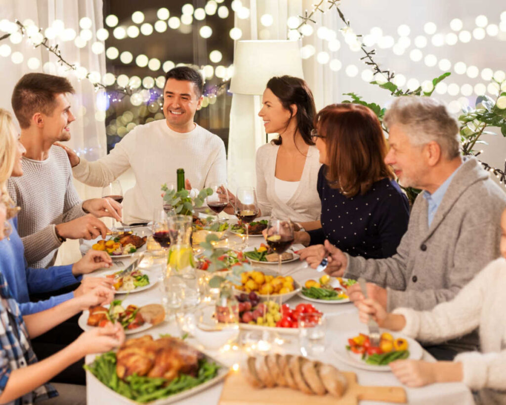 A diverse group of family and friends smiling and talking while gathered around a dining table for a festive indoor meal. The table is laden with food, including a whole roast chicken or turkey, sliced bread, and side dishes. String lights and warm bokeh lights decorate the background.