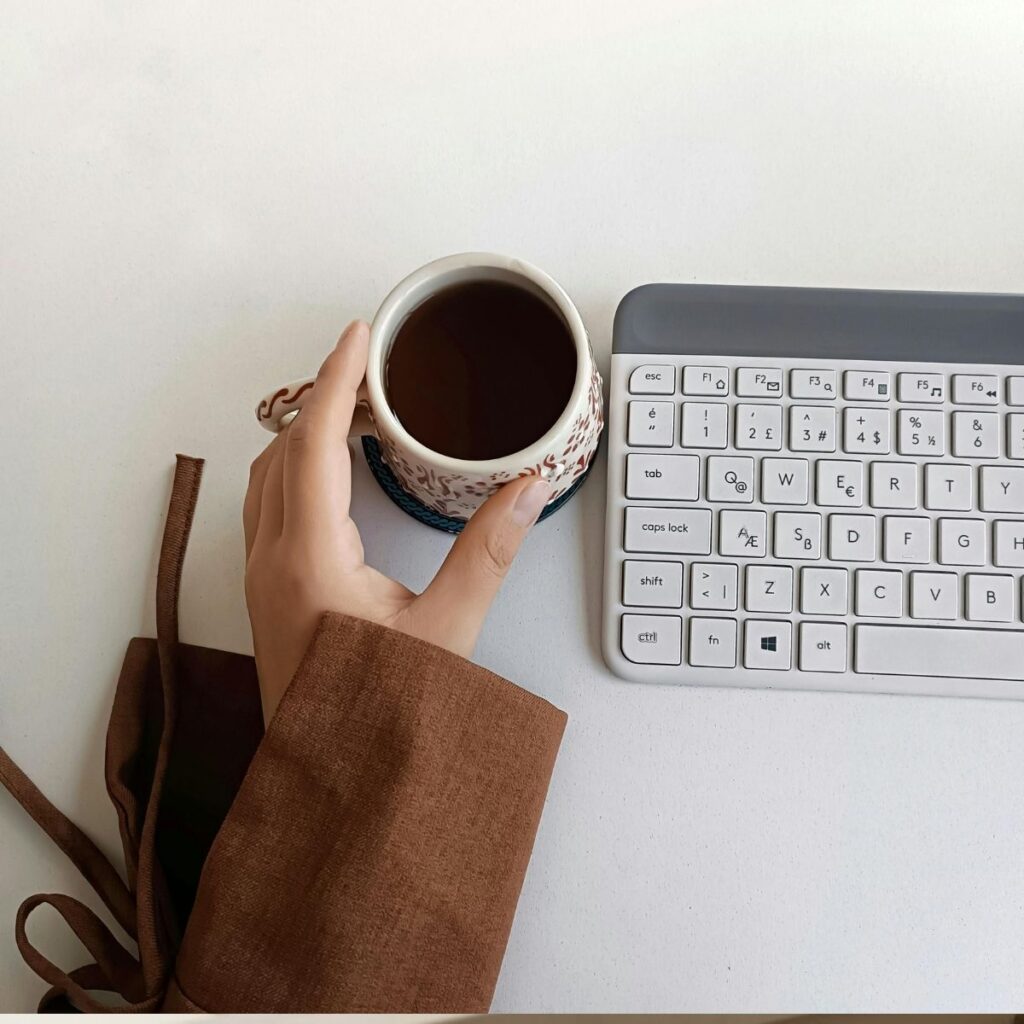 Hand in a brown sleeve holding a patterned mug of tea or coffee next to a small white and gray computer keyboard.