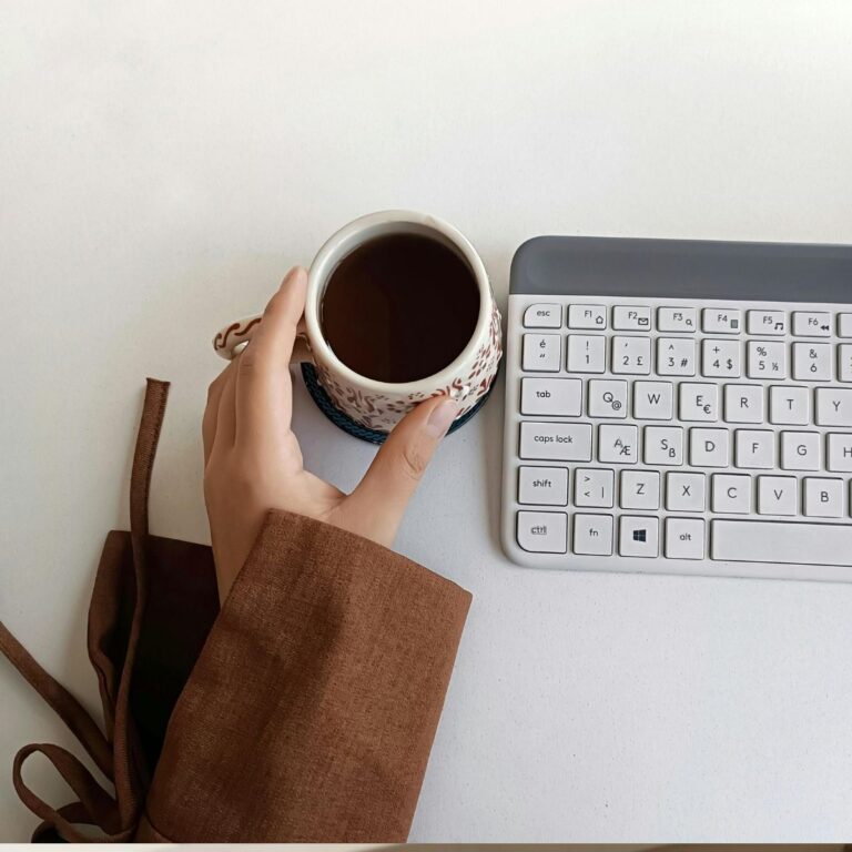 Hand in a brown sleeve holding a patterned mug of tea or coffee next to a small white and gray computer keyboard.