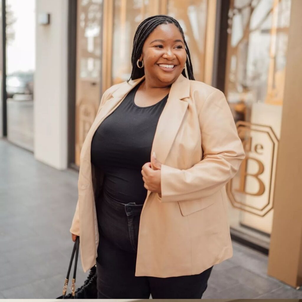 Woman in a beige leather blazer smiling on a city sidewalk.