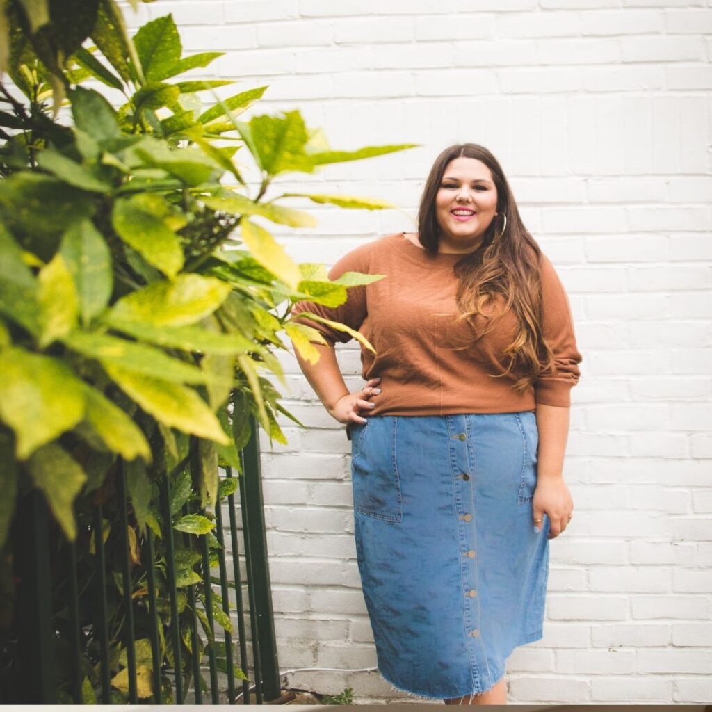 Woman smiling in a denim skirt and brown sweater against a white wall.