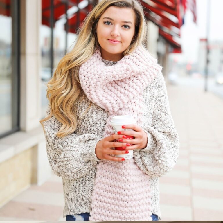 Young blonde woman in a gray chunky sweater and oversized pale pink knit scarf holds a coffee cup on a city street.
