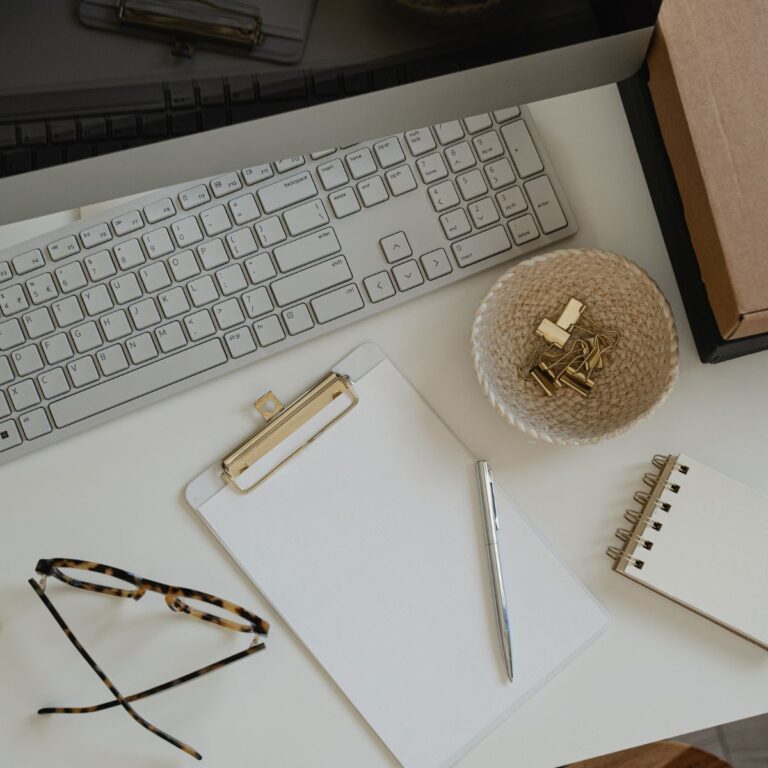 Overhead view of a white desk with a keyboard, clipboard and pen, spiral notebook, eyeglasses, and a bowl of gold binder clips.