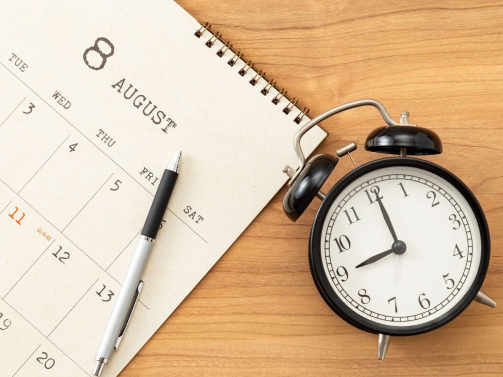 August calendar page with a pen beside a black alarm clock on a wooden surface.