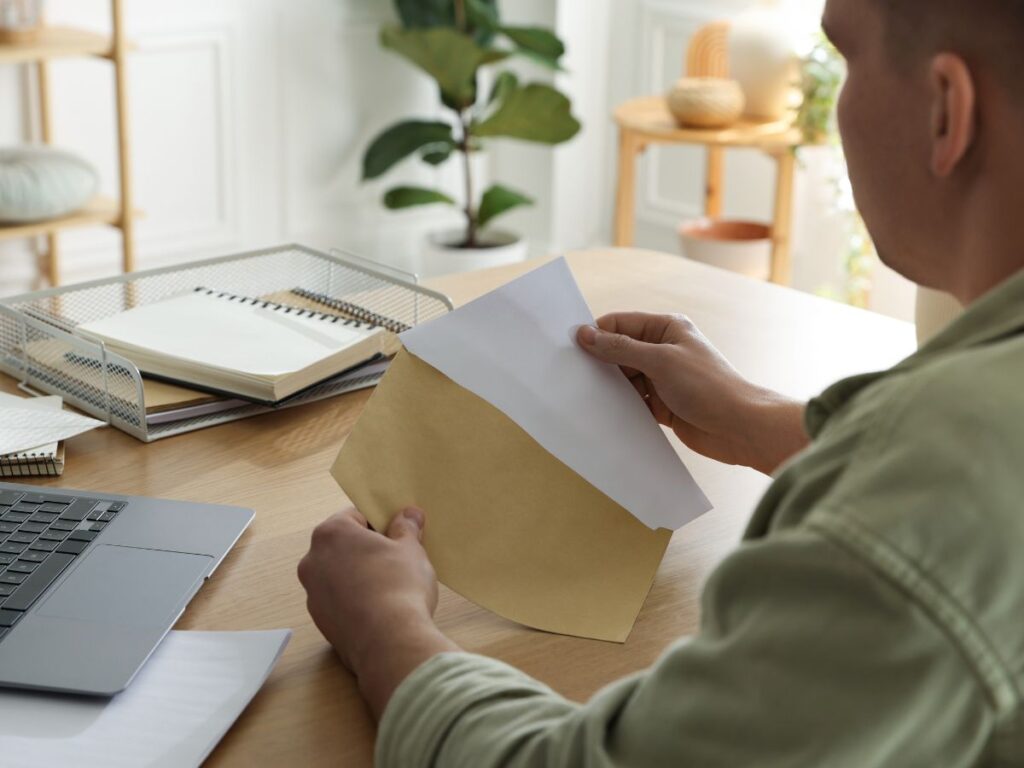 Man sitting at a wooden desk opening a paper envelope, with a laptop and stacked documents nearby.