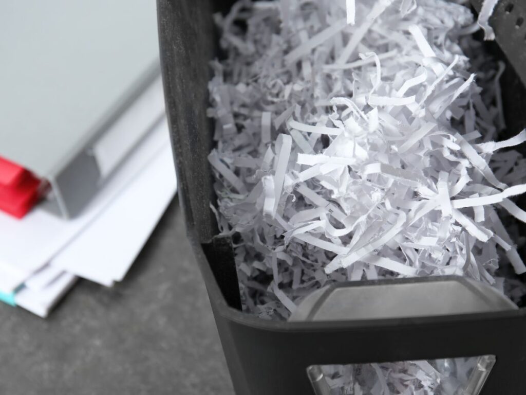 Close-up of a paper shredder bin filled with shredded white paper, with a stack of documents nearby.