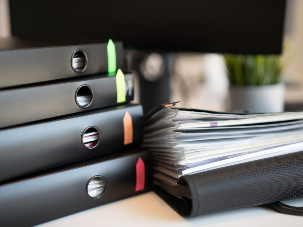 Stack of black ring binders with colorful tabs beside an accordion file folder filled with papers on a desk.