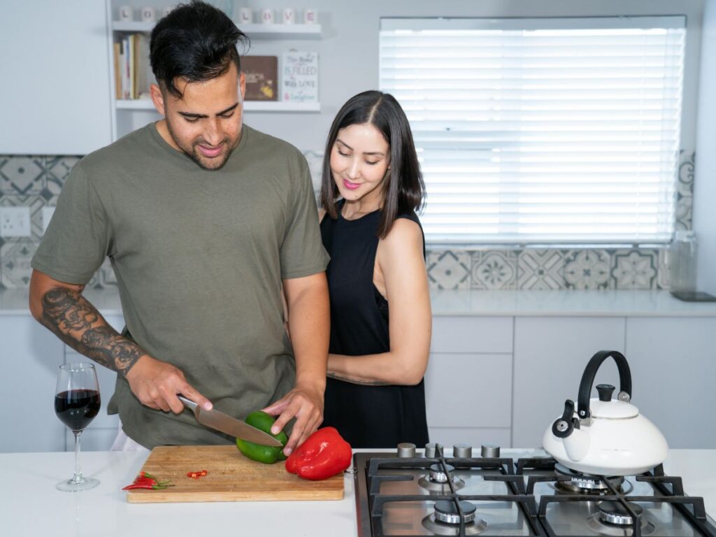 A couple cooking together in a bright kitchen, chopping bell peppers on a cutting board beside a glass of wine.