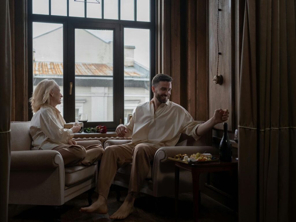 A couple relaxing by a large window, sitting in armchairs and sharing wine and snacks in a cozy, softly lit room.