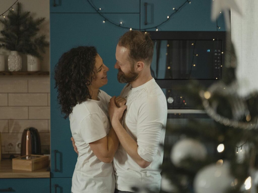 A couple standing close together in a cozy kitchen, smiling warmly at each other. Soft string lights and a decorated Christmas tree in the foreground add a festive, intimate atmosphere.