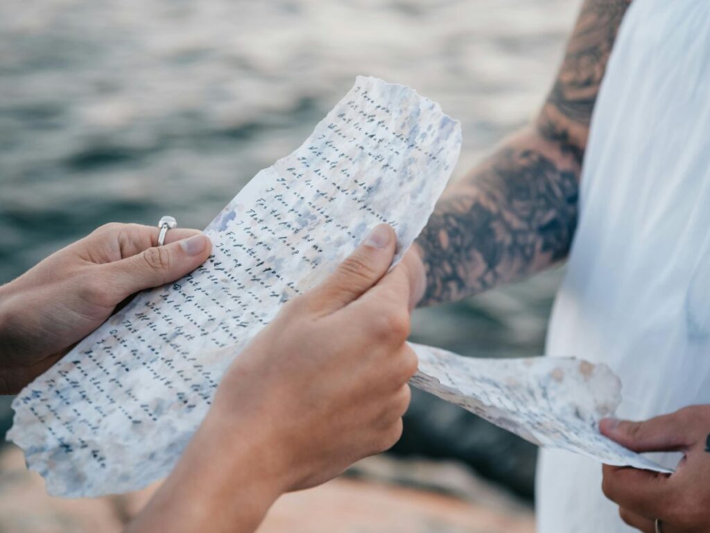 Close-up of two people holding and reading handwritten vows on textured paper by the water, one with a tattooed arm.