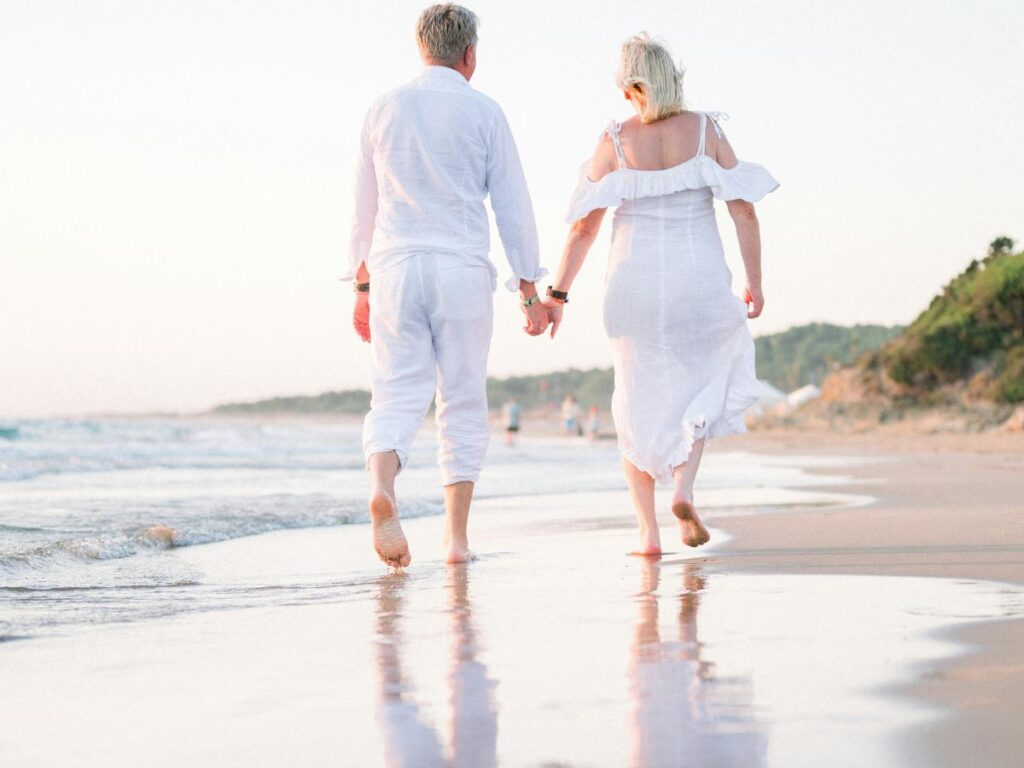 An older couple dressed in white walking hand in hand along the shoreline at sunset, their reflections visible in the wet sand.