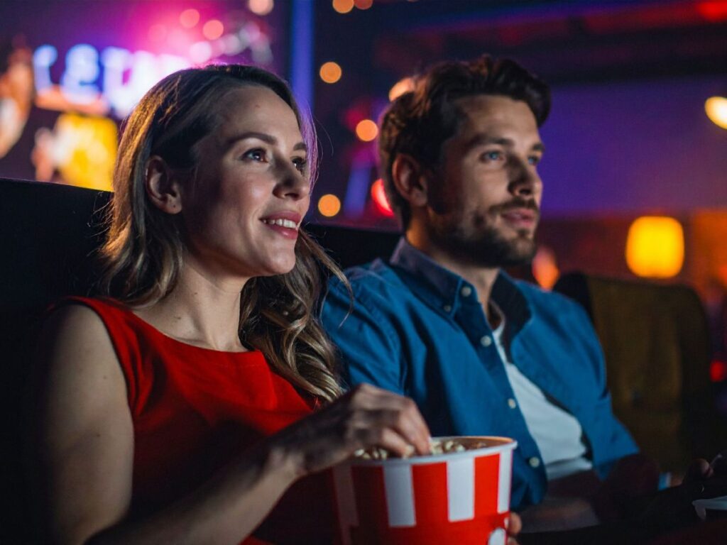 A couple sitting in a movie theater, smiling and sharing popcorn while watching a film.
