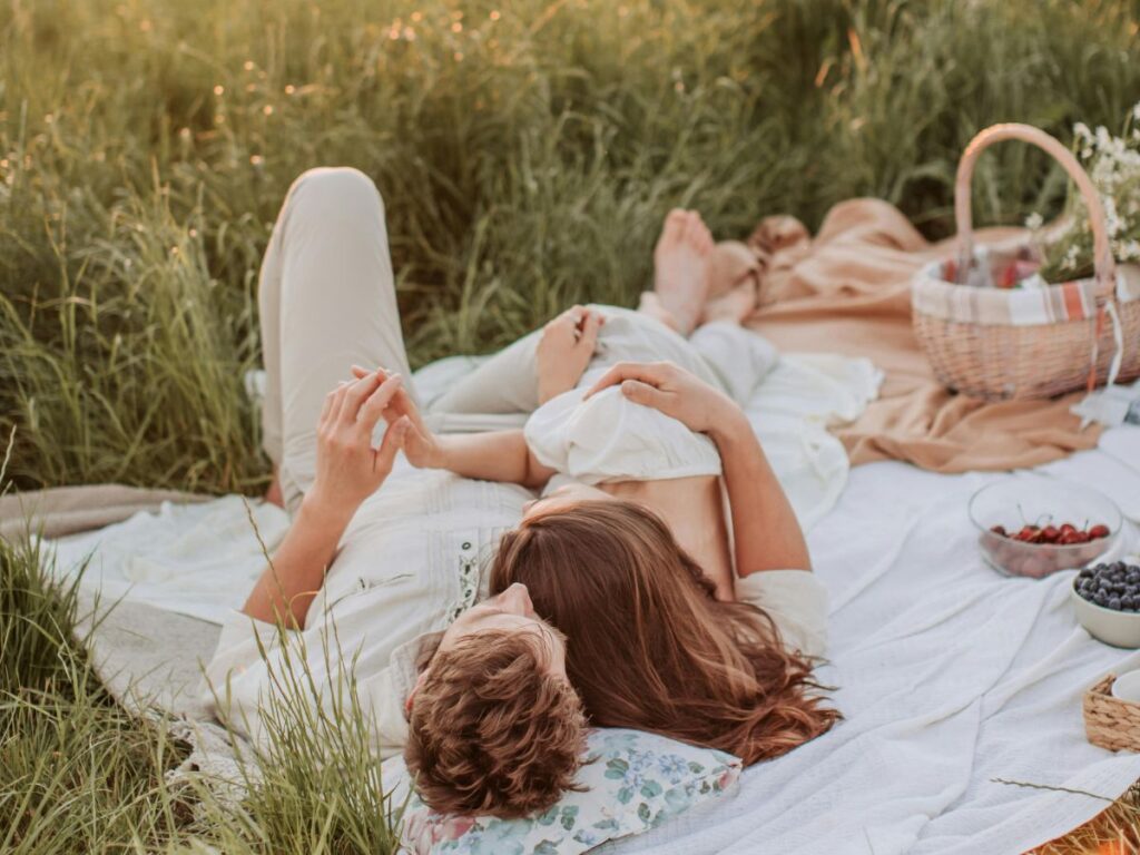 A couple lying on a picnic blanket in a grassy field at sunset, relaxing together with a basket and fruit nearby.