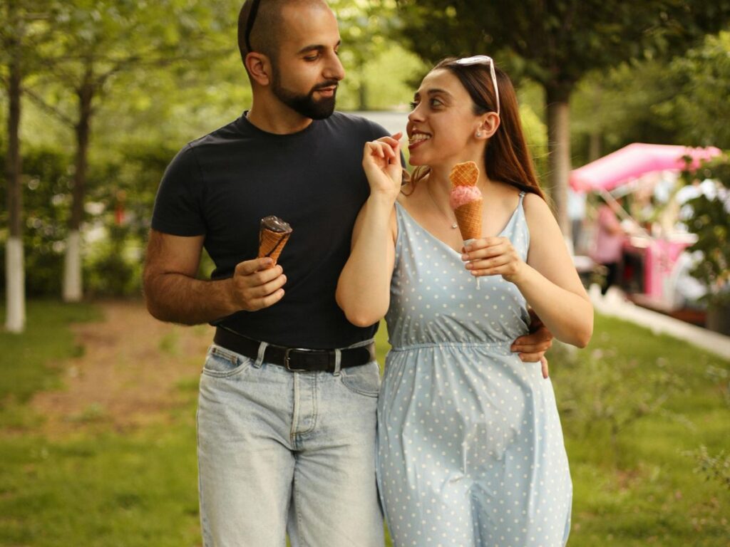 A couple walking arm in arm through a green park, smiling at each other while enjoying ice cream cones on a sunny day.